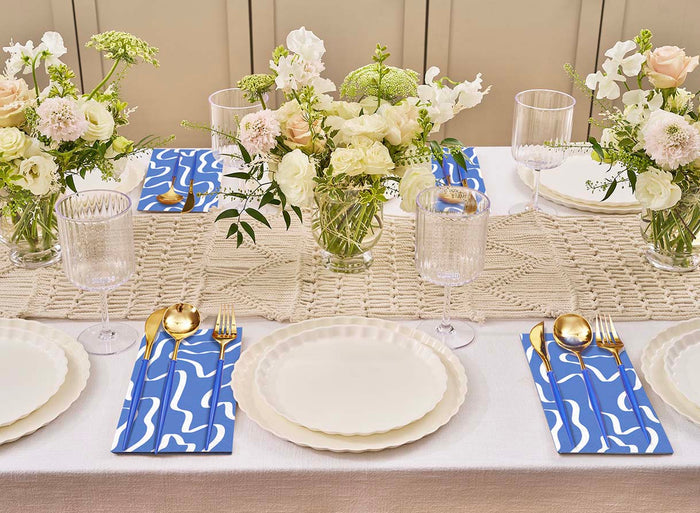 The Sophistiplate Eco White & Blue Table Setting features eco-friendly tableware for four, gold cutlery on blue abstract-pattern napkins, clear glasses, and vases with white and pale pink flowers on a crocheted runner.
