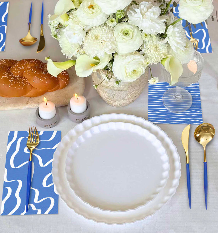 The Sophistiplate Eco White & Blue Table Setting features eco-friendly tableware, blue-and-white abstract napkins, gold and blue cutlery, a floral centerpiece, candles, challah bread, all atop a white tablecloth.