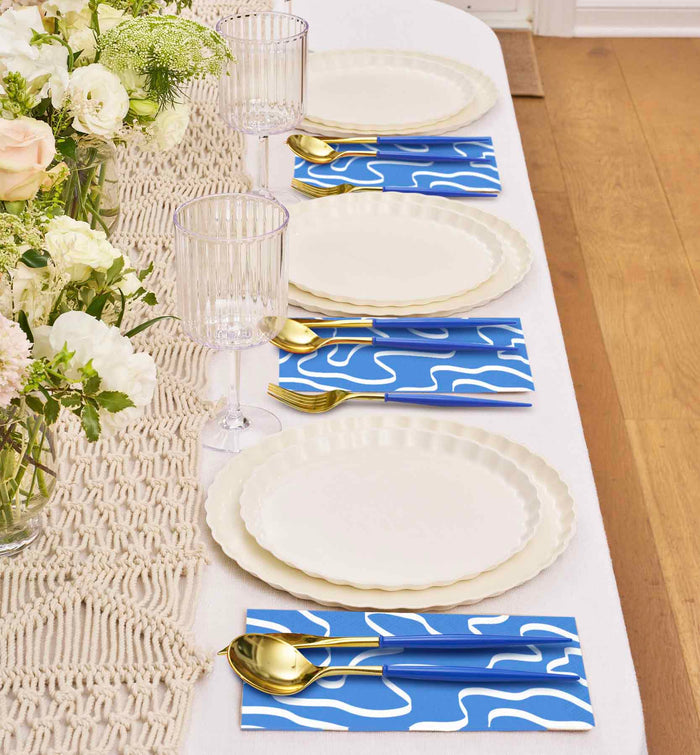The Eco White & Blue Table Setting by Sophistiplate features cream plates, gold cutlery, blue napkins with white patterns, ribbed glass goblets, and a macrame runner adorned with elegant floral arrangements.