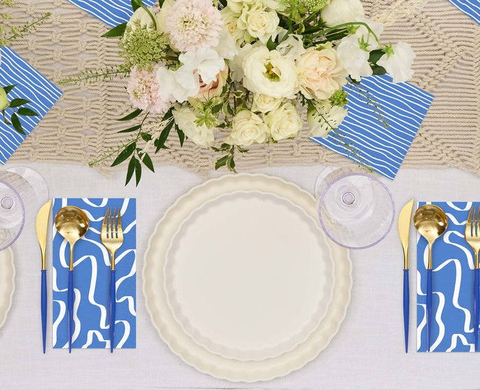 The Eco White & Blue Table Setting by Sophistiplate features eco-friendly scalloped white plates, gold cutlery, blue and white abstract napkins, clear glasses, and a centerpiece of white and pale pink flowers on a woven beige runner.