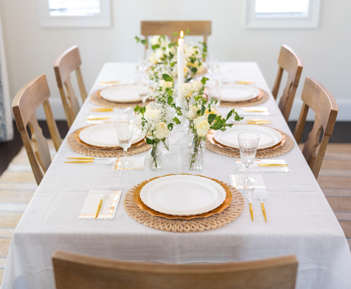 The Sophistiplate LLC Eco White & Gold Table Setting seats six with designer disposable plates, gold utensils, woven placemats, clear glasses, white napkins, floral centerpieces of white and yellow blooms, and a lit candle centerpiece.