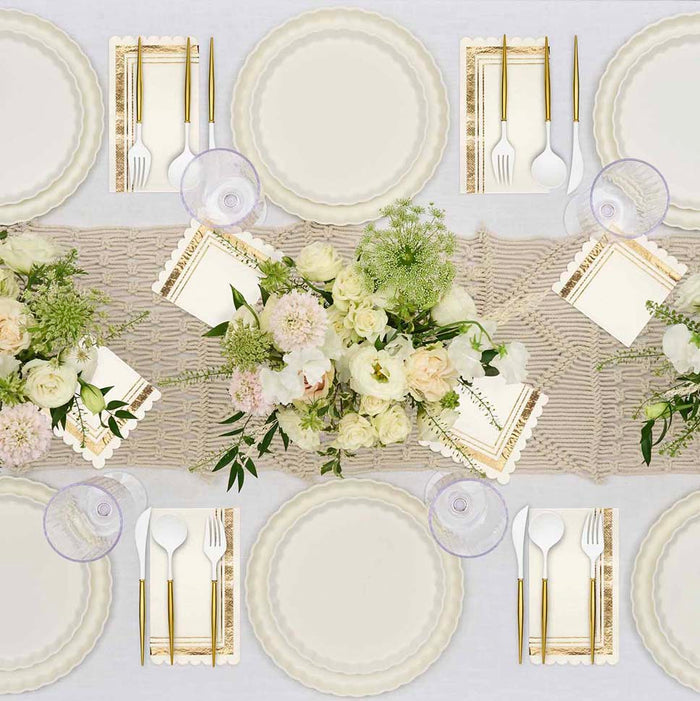 The Eco White & Gold Table Setting by Sophistiplate LLC features cream eco-friendly plates, gold cutlery, clear glasses, and decorative napkins on a lace runner with white and blush floral centerpieces for a formal table.