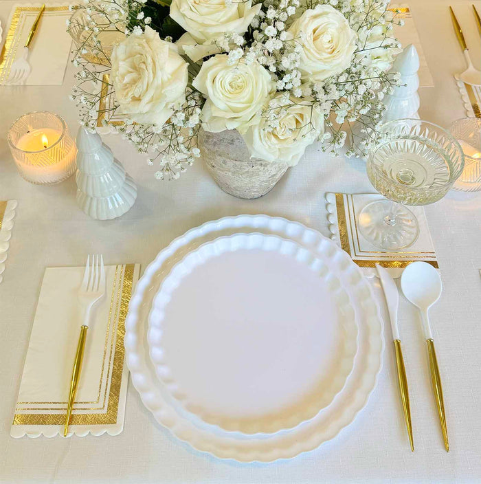 The Eco White & Gold Table Setting by Sophistiplate LLC features eco-friendly plates, gold-accented cutlery, napkins, candles, glassware, and a centerpiece of white roses and baby's breath in a vase.