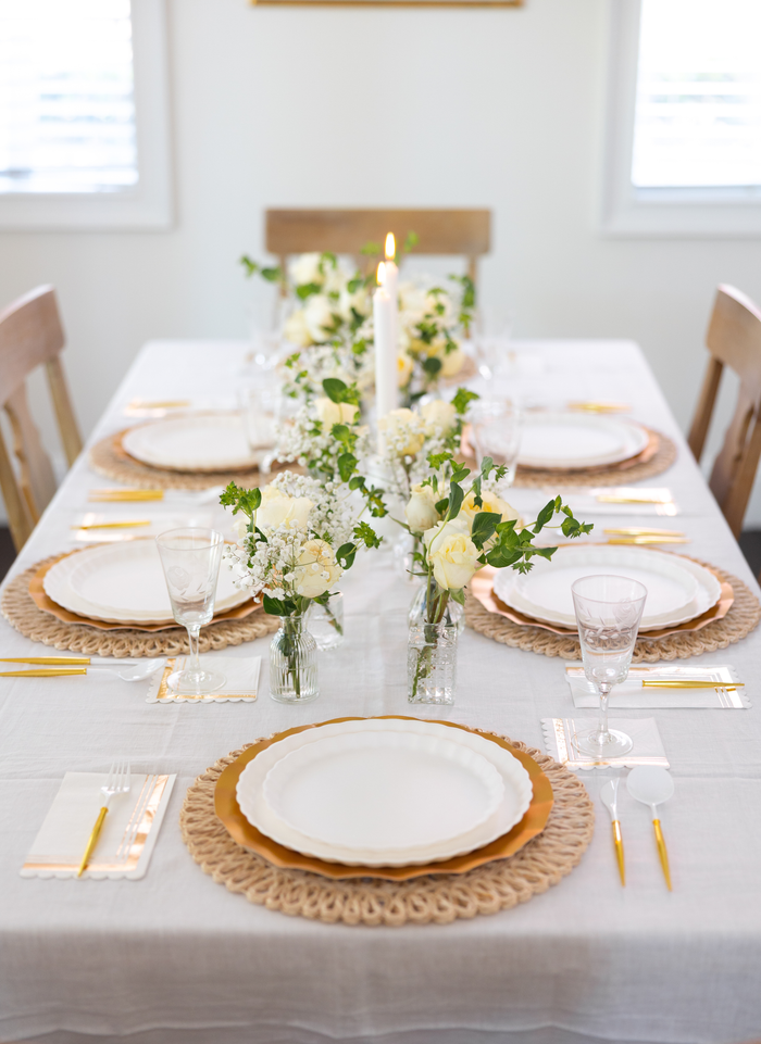 The Eco White & Gold Table Setting by Sophistiplate LLC includes designer disposable plates, gold chargers, gold flatware, crystal glasses, and white napkins for six. The centerpiece has white flowers, greenery, and a single lit candle.