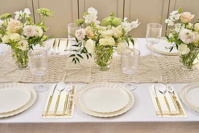 The Eco White & Gold Table Setting by Sophistiplate LLC features eco-friendly plates, gold cutlery, clear glasses, and elegant cream and pale pink floral centerpieces on a textured beige table runner.