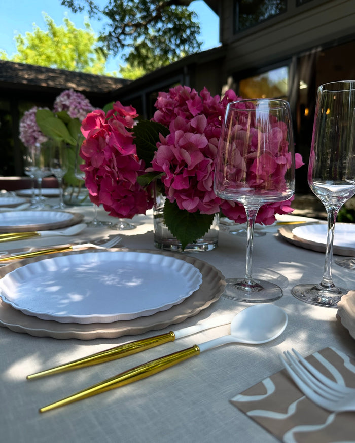 A close-up of the Sophistiplate Eco-Friendly Table Setting-Sand, featuring gold and white cutlery, scalloped white plates, wine glasses, and pink hydrangeas in a glass vase on an outdoor table with trees and a house in the background.
