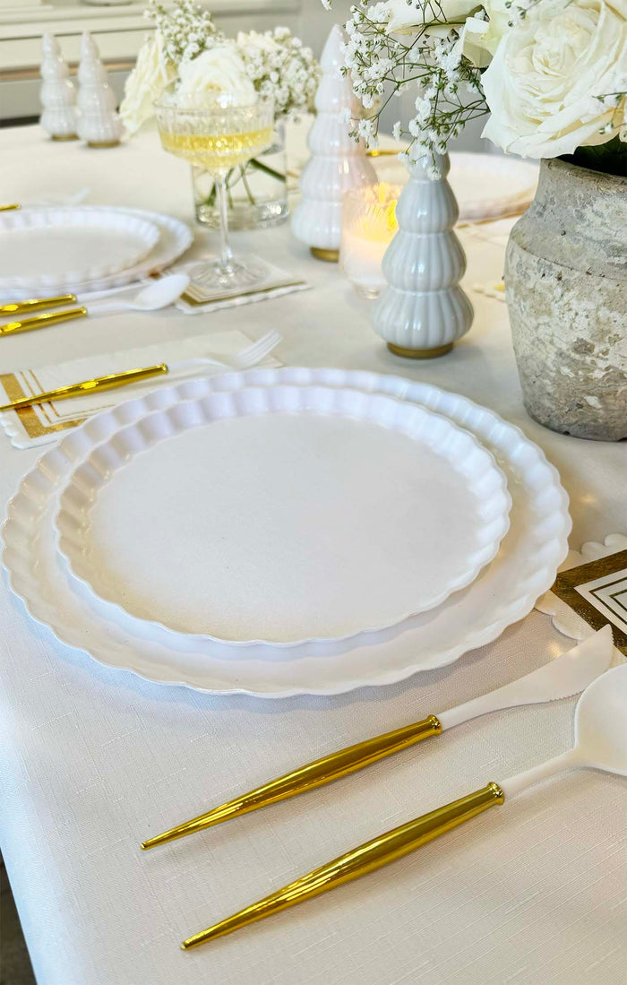 A close-up of the Sophistiplate Eco White & Gold Table Setting, featuring white scalloped biodegradable bamboo plates, gold-colored cutlery, white napkins, and elegant floral ceramic centerpieces on a white tablecloth.
