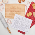 A handwritten gingerbread cookie recipe sits beside the Maple Engraved Recipe Arched Artisan Maple Board (12 x 9"), with baking essentials—rolling pin, eggs, flour, cookie cutters—and a red polka-dot cloth on a white surface.