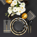 The Sophistiplate Everyday Black Table Setting features elegant gold-rimmed paper plates, gold and black cutlery, striped napkins, golden cups, and a white floral centerpiece on a black tablecloth.