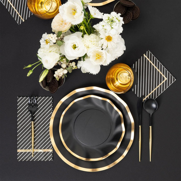The Sophistiplate Everyday Black Table Setting features elegant gold-rimmed paper plates, gold and black cutlery, striped napkins, golden cups, and a white floral centerpiece on a black tablecloth.