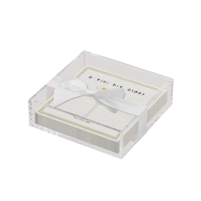 The Sophistiplate Everyday Cocktail Napkin with Napkin Tray features a clear acrylic tray, ideal with your ceramic napkin holder, containing white napkins with gold borders. The top napkin says "a tiny bit older" in lowercase, accented by a gold triangle.