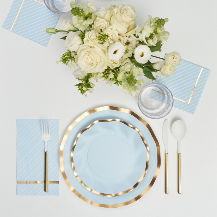The Sophistiplate Everyday Sky Blue Table Setting includes blue and gold-rimmed designer disposable plates, gold-handled cutlery, a glass, a blue striped napkin, and a bouquet of white and cream flowers on a white background.