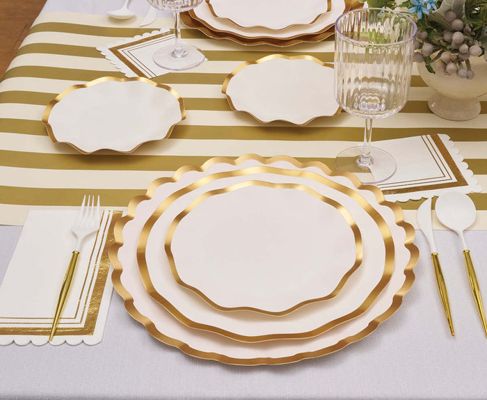 A table is elegantly arranged with white and gold scalloped plates, gold utensils, a wine glass, a napkin, and a striped gold and white table runner. In the corner rests a small floral arrangement, beautifully complemented by Sophistiplate’s Everyday White Appetizer/Dessert Bowls (8pk), which feature refined gold trim to enhance the sophisticated setting.