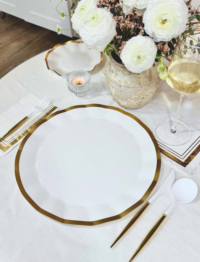 Elegant table setting with Sophistiplate Everyday White Charger Plates (8pk), gold cutlery, a wine glass, white flowers in a vase, a candle, and a neatly folded napkin on a white tablecloth.