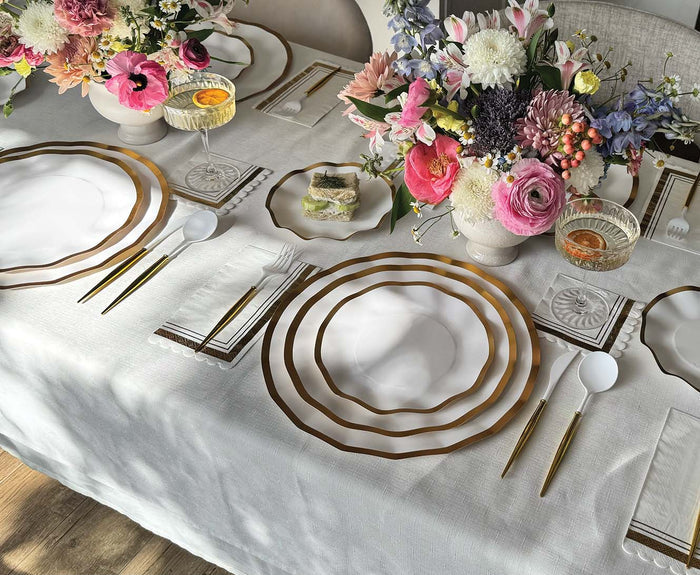 Set an elegant table with Sophistiplate’s Everyday White Salad Plates (8-pack), featuring scalloped edges, paired with gold flatware, glassware, floral centerpieces, and a slice of dessert on a small plate—all arranged atop a white tablecloth in natural light.