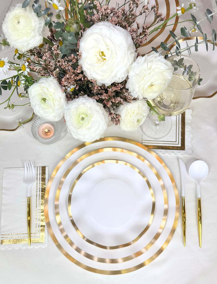 Sophistiplate Everyday White Charger Plates (8pk) add elegance to your table, paired with gold and white cutlery, a wine glass, candle, and a white ranunculus & greenery centerpiece on a crisp white tablecloth.