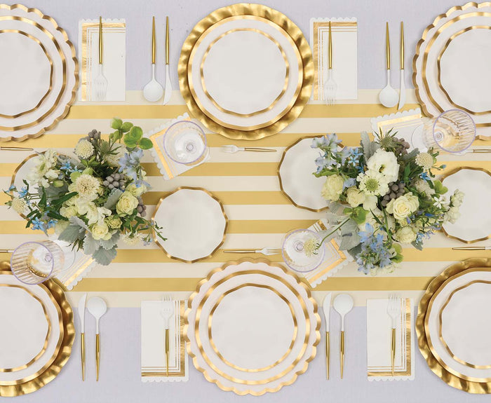 Top-down view of an elegant table set featuring Everyday White Dinner Plates/8pk by Sophistiplate, adorned with gold trim and scalloped edges, paired with gold utensils, a striped gold and white table runner, white napkins, and two floral centerpieces with green and white flowers.