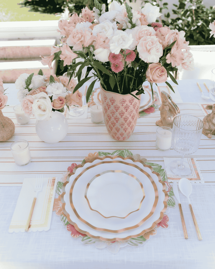 An elegant table setting features Sophistiplate Everyday White Dinner Plates (8pk), gold utensils, white napkins, glassware, candles, and vases of pink and white flowers arranged on a striped table runner over a white tablecloth.
