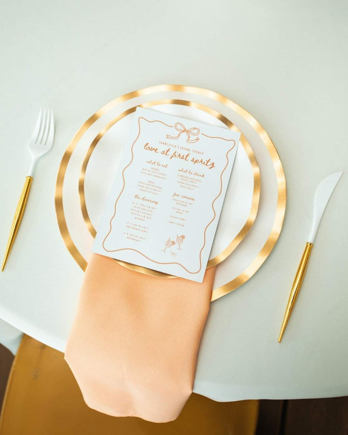 An elegant table setting featuring Sophistiplate’s Everyday White Dinner Plates (8pk), paired with gold-accented cutlery, a plate charger, a peach napkin, and a white menu card on a round table draped with a white tablecloth.