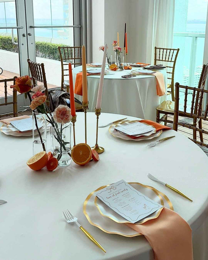 Round tables set for an event feature menus, gold cutlery, pink and orange candles, fresh flowers, and halved citrus fruits as vibrant centerpieces. Each place setting is elevated with chic Everyday White Dinner Plates from Sophistiplate (8-pack), all beautifully arranged near expansive windows overlooking the ocean.