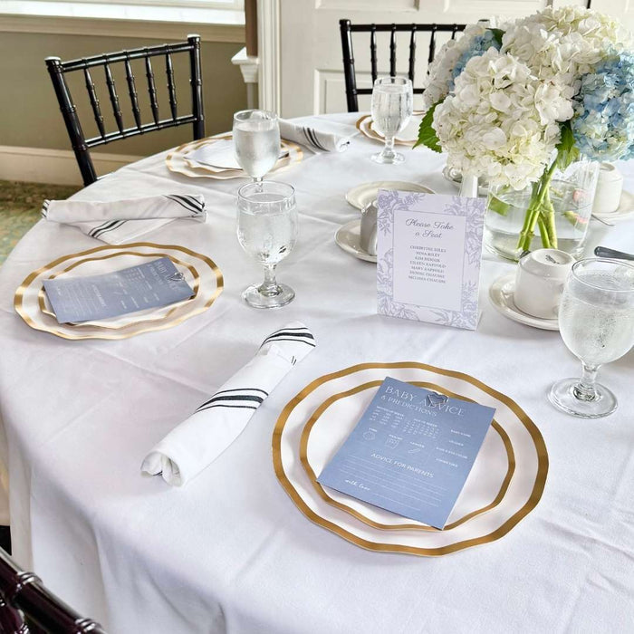 A round table set with Sophistiplate Everyday White Dinner Plates (8-pack), elegantly accented by folded napkins, water glasses, and a floral centerpiece of white and blue blooms. Menus and an event card are also displayed on the table.