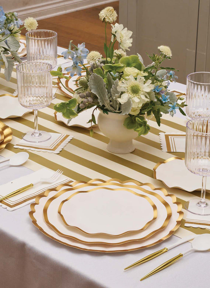 A table set with gold flatware, crystal glasses, white napkins, and a centerpiece of white and green flowers on a gold-striped runner offers classic elegance—made even more convenient with the addition of disposable plates featuring gold trim. For an extra touch of sophistication, add the Everyday White Salad Plates/8pk by Sophistiplate; their scalloped edges bring delicate charm that elevates the whole setting.