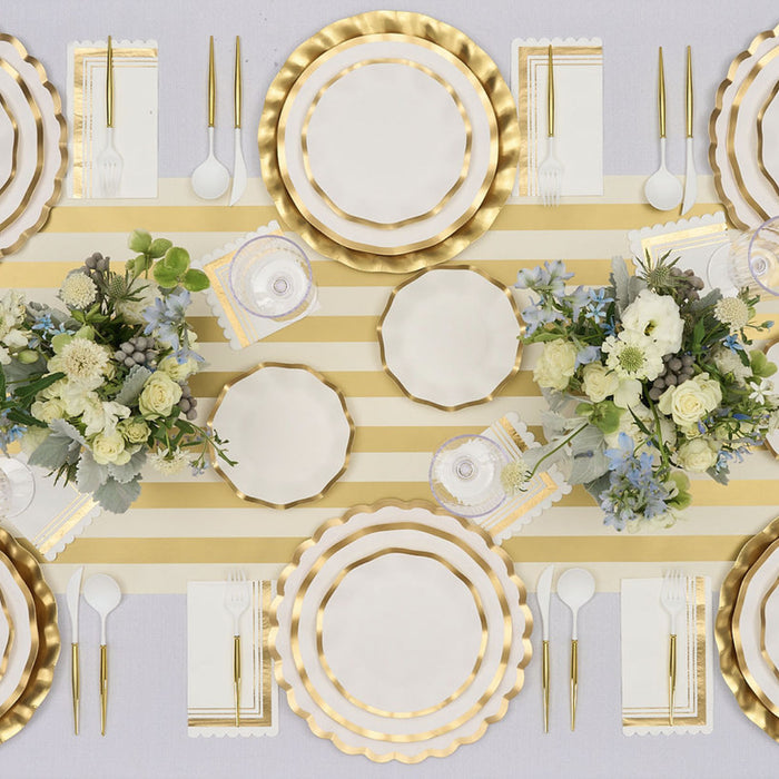 The Everyday White Table Setting by Sophistiplate LLC seats eight and includes gold-rimmed plates, gold cutlery, white napkins, a gold and white striped runner, and floral centerpieces in white, green, and blue.