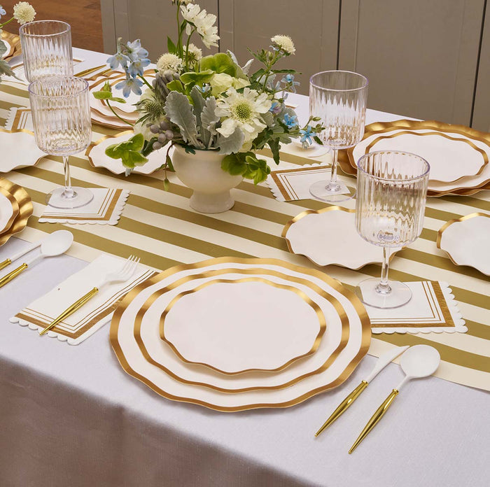 The Gilded Honey Table Setting by Sophistiplate LLC features designer gold-rimmed white disposable plates, gold and white cutlery, crystal glasses, a striped gold and white table runner, and a white and blue floral centerpiece.