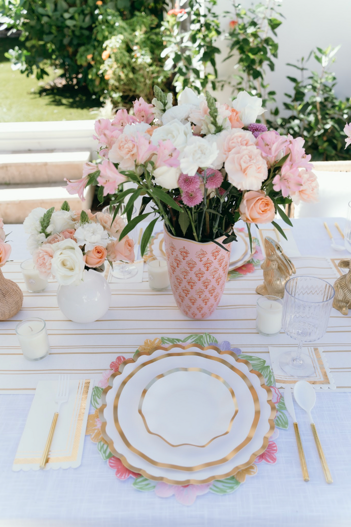 Elegant outdoor table setting features a floral centerpiece of pink, peach, and white blooms, surrounded by glowing white candles and gold utensils. The crisp white tablecloth is layered with Sophistiplate’s Everyday White Salad Plates/8pk—scalloped-edged paper plates offering both style and convenience for your gathering. Garden greenery in the background adds to the charming atmosphere.