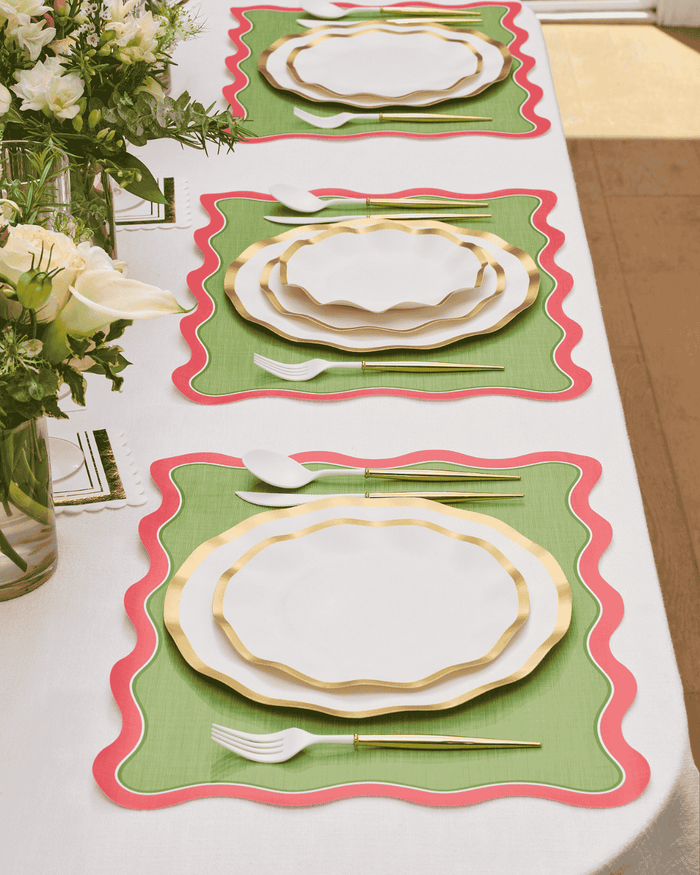 A table set for three features Sophistiplate’s Everyday White Dinner Plates, paired with gold flatware and green placemats trimmed in red. White flowers in vases adorn the white tablecloth, while natural light illuminates the setting.