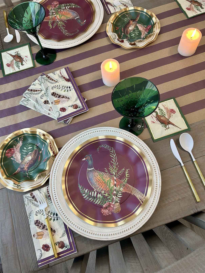 The Feather Grove Table Setting by Sophistiplate features a striped purple and beige tablecloth, pheasant-themed plates, green goblets, gold-handled cutlery, candles, and napkins with floral and bird designs for a perfect fall tablescape.