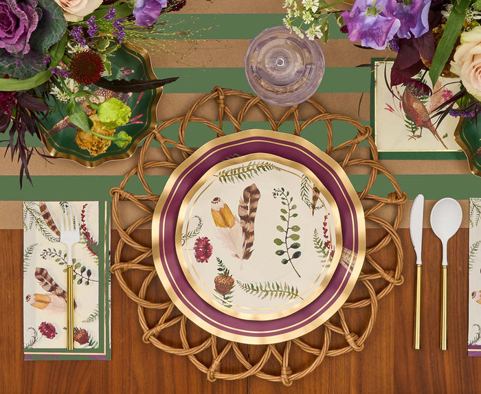 The Feather Grove Table Setting by Sophistiplate features a woven charger, botanical feather plate, gold-trimmed utensils, crystal glass, and floral napkins, accented with purple and cream blooms for an elegant fall tablescape.