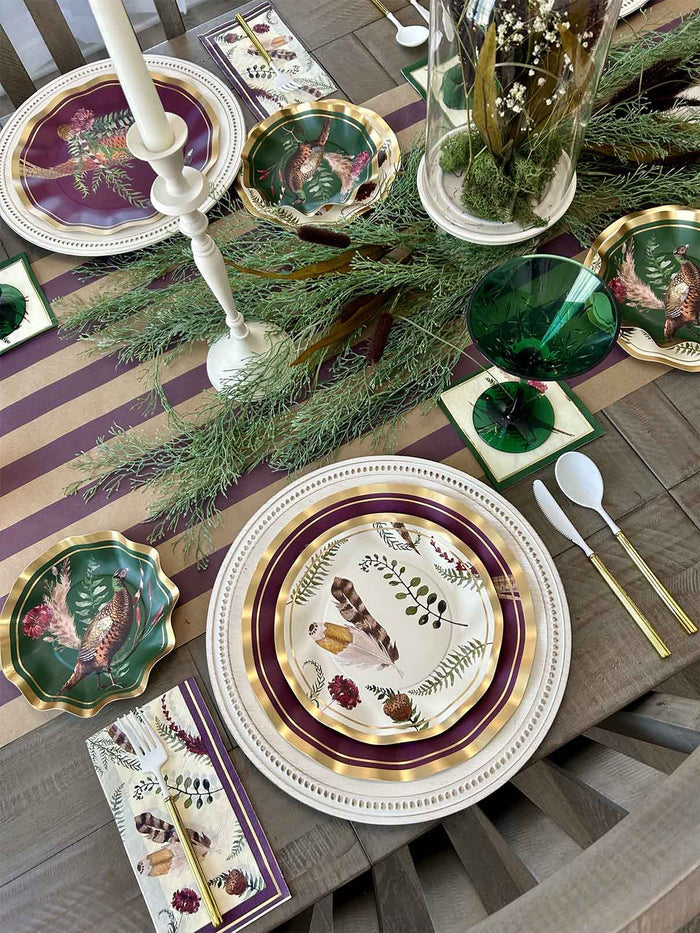 Create a festive fall tablescape with the Sophistiplate Feather Grove Table Setting—elegant bird and botanical plates, napkins, green glassware, white candlesticks, green garland, and a purple-and-gold striped runner on wood.