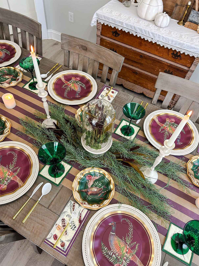 A festive dining table is set with maroon and gold plates featuring a bird motif, green glasses, candles, evergreen branches, and a striped table runner. Sophistiplate’s Feather Grove Appetizer/Dessert Bowls (8pk) are artfully arranged at each place setting, adding an extra touch of autumn charm. In the background, a wooden sideboard decorated with pumpkins and draped with a lace cloth completes the cozy scene.