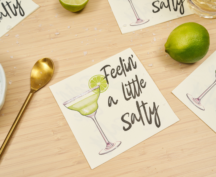 Sugarplum’s Feeling a little Salty Cocktail Napkins (26pk) feature a margarita illustration and playful text, shown on wood with a gold spoon, whole lime, lime wedge, and salt for a summery vibe.