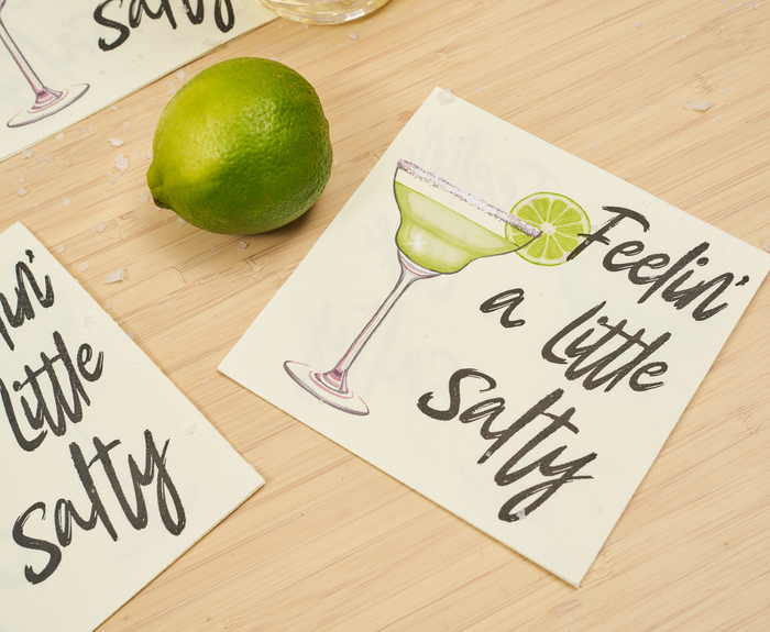 A lime rests on a light wooden surface sprinkled with salt, alongside a Sugarplum “Feeling a little Salty Cocktail Napkin” (26pk) featuring a margarita sketch—perfect for playful summer gatherings.
