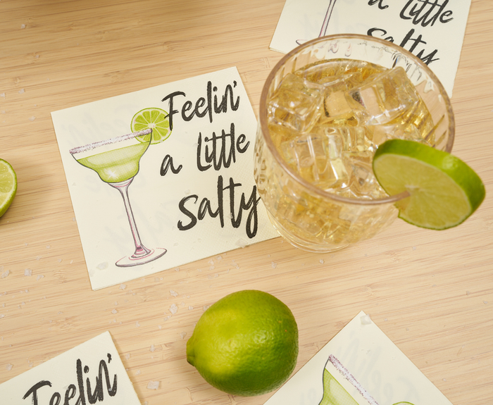 A glass of cocktail with ice and lime rests on Sugarplum’s Feeling a little Salty Cocktail Napkins (26pk), while a whole lime and other playful party napkins decorate the wooden surface.