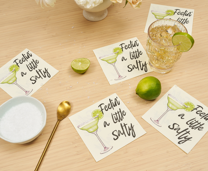 A wooden tabletop displays Sugarplum’s Feeling a little Salty Cocktail Napkins (26pk) with margarita art, a glass of iced drink with lime, whole and sliced limes, a bowl of salt, and a gold spoon.