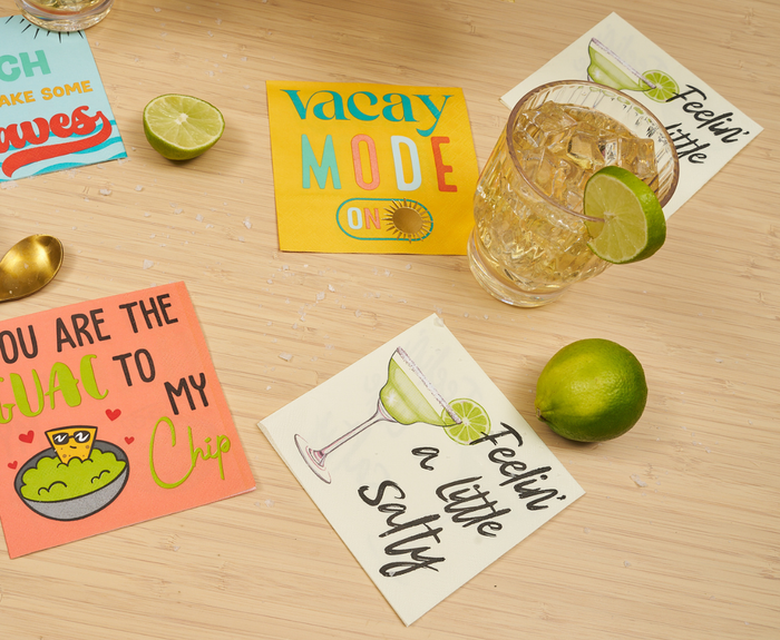 A tabletop with Sugarplum’s "Feeling a little Salty" cocktail napkins (26pk), a glass of iced drink with a lime wedge, two limes (one whole, one sliced), a gold spoon, and scattered salt.