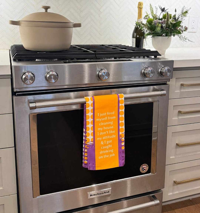 A beige pot, champagne bottle, and flower vase sit on the stovetop above an oven with the Drinks On Me "Fired Dish Towel," which says: I just fired myself from cleaning my house—ideal kitchen humor for a housewarming gift.