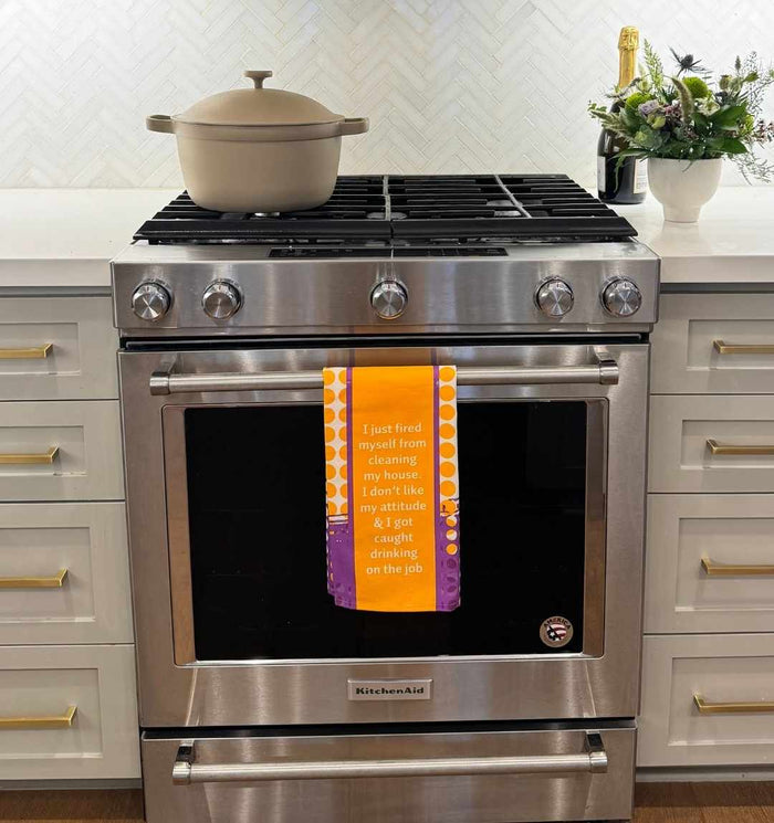 The Fired Dish Towel by Drinks On Me, featuring the quote "I just fired myself from cleaning my house…", hangs on a modern stainless steel oven—funny kitchen décor and a unique housewarming gift. A beige pot and fresh flowers complete the scene.