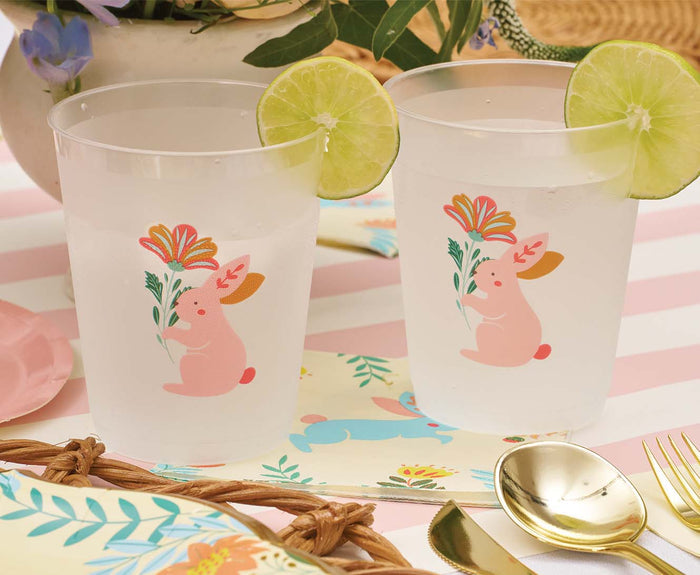 The Ultimate Easter Tablescape for 16: Folklore Easter by Sophistiplate LLC features bunny and flower cups, festive napkins, gold cutlery, disposable plates, and green leaves for a cheerful spring celebration.