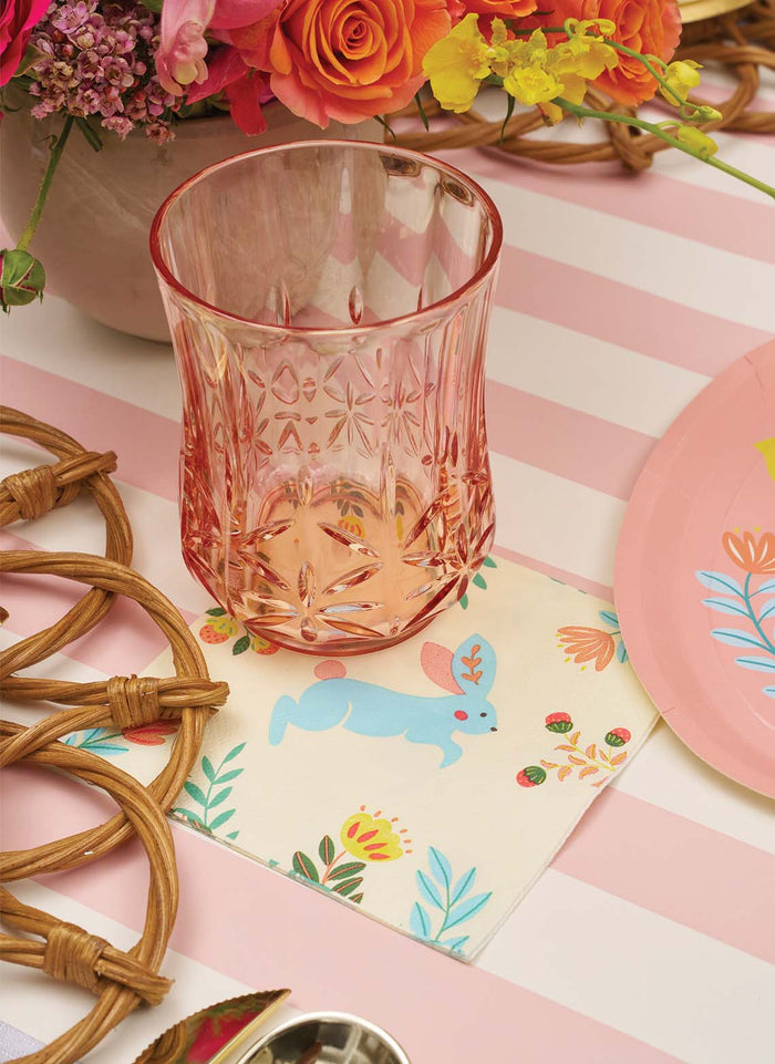 A pink patterned glass rests on Sophistiplate Folklore Easter Cocktail Napkins (20pk) featuring a blue bunny and flowers, beside a pink plate, rattan placemats, and a floral centerpiece atop a pink striped tablecloth.