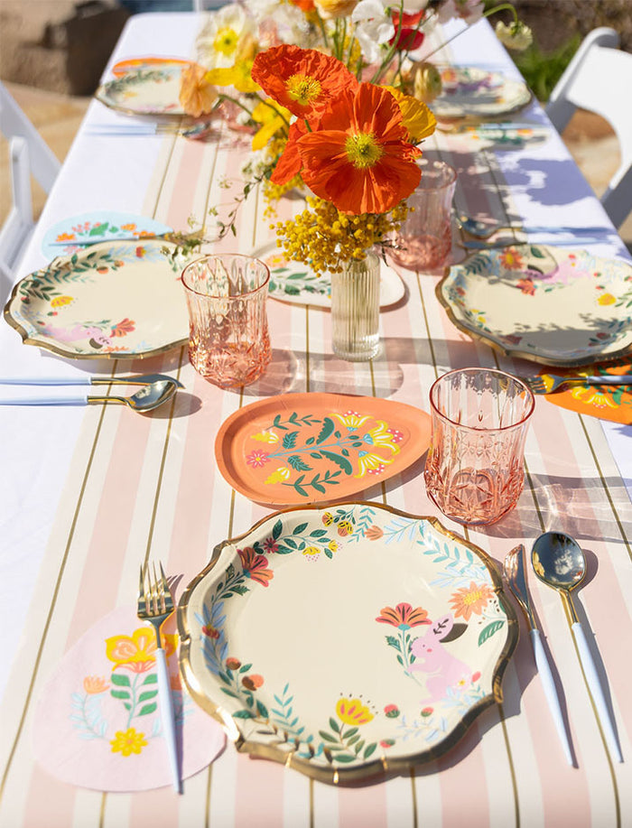 The Ultimate Easter Tablescape for 16: Folklore Easter by Sophistiplate LLC features designer plates, pink glasses, gold utensils, and vibrant flowers on a striped cloth for a cheerful spring setting.