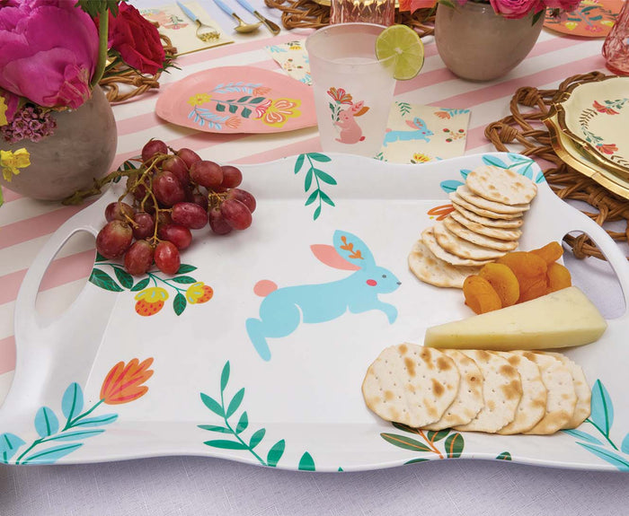 The Sophistiplate LLC Ultimate Easter Tablescape for 16: Folklore Easter features a white tray with a blue bunny and floral design, beautifully presenting grapes, crackers, cheese, and apricots among flowers and festive decor.
