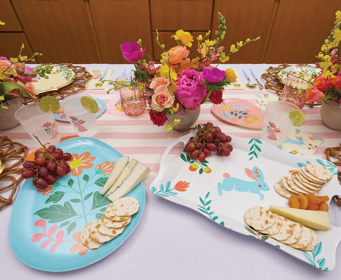 A vibrant spring table features floral plates, a Sophistiplate Folklore Easter Melamine Tray with cheese, crackers, grapes, and apricots, bright flower arrangements, drinks with lime slices, and a pink and white striped runner.