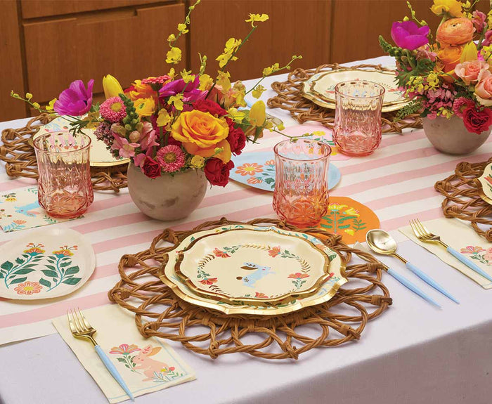 The Folklore Easter Table Setting by Sophistiplate features floral centerpieces, bird-themed designer plates, woven placemats, pastel cutlery, pink glasses, a striped table runner, and floral coasters for a vibrant spring party.