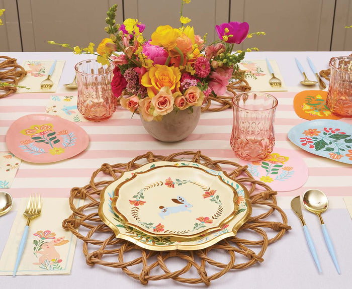 The Ultimate Easter Tablescape for 16: Folklore Easter by Sophistiplate LLC features pastel designer plates, gold cutlery, pink glasses, and a bouquet of yellow and pink flowers on a striped tablecloth.