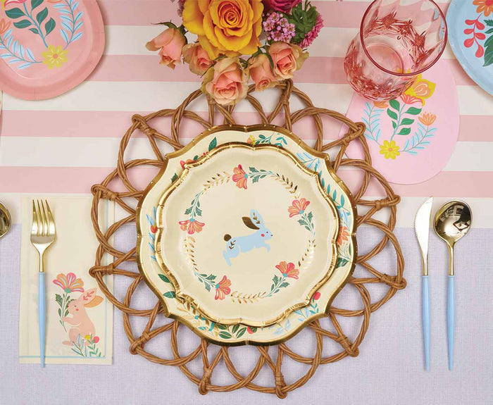 The Ultimate Easter Tablescape for 16: Folklore Easter by Sophistiplate LLC features designer bunny plates, gold-rimmed cutlery, pink glasses, a striped runner, and a yellow and pink floral centerpiece for a charming pastel-themed setting.
