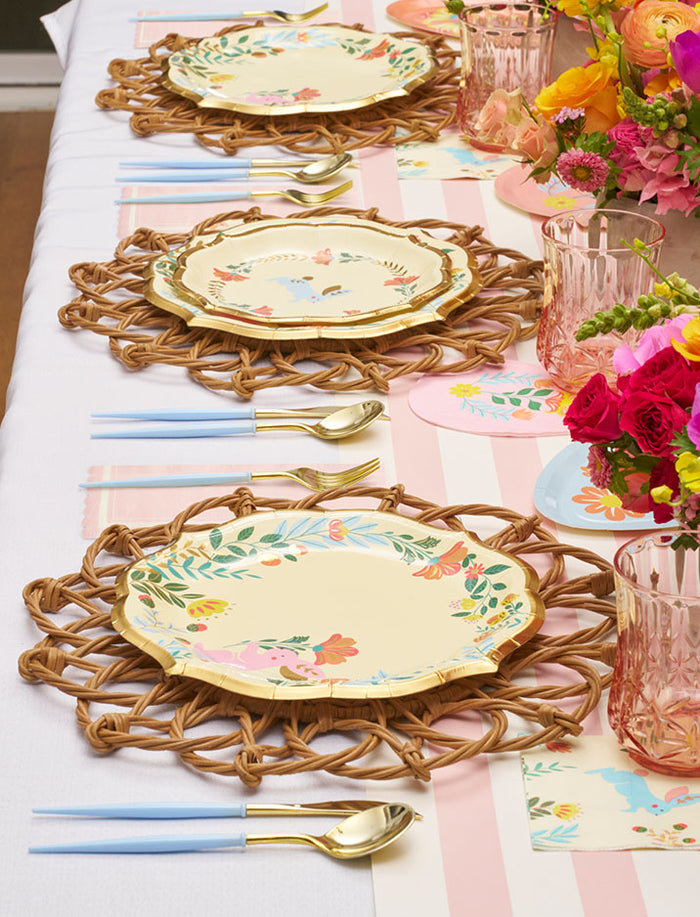 The Ultimate Easter Tablescape for 16: Folklore Easter by Sophistiplate LLC features woven placemats, designer plates, gold cutlery with blue handles, pink glasses, and a vibrant floral centerpiece on a white and pink striped tablecloth.